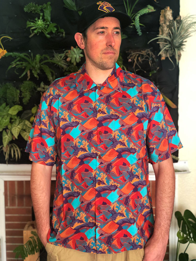 Man in colorful Hawaiian shirt with orange and blue design, standing in front of a wall full of plants