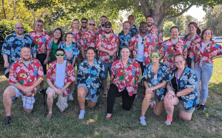 A photo of a group of people wearing Hawaiian shirts. Five people in front are kneeling for the photo.