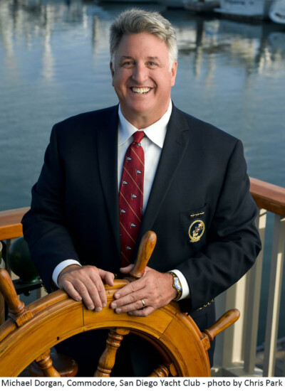 "Thank you to April and the team at Shop4Ties for helping to accommodate our design, make and deliver on time the exact tie we wanted for our 2018 San Diego Yacht Club Board of Directors uniform — they look great and on a relative short notice!" - Michael Dorgan, Commodore, San Diego Yacht Club Photo credit: Christopher Park Man stands at a ship steering wheel, smiling, wearing a custom tie and a suit jacket.