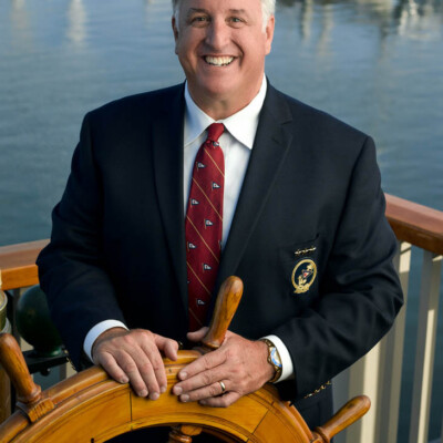 "Thank you to April and the team at Shop4Ties for helping to accommodate our design, make and deliver on time the exact tie we wanted for our 2018 San Diego Yacht Club Board of Directors uniform — they look great and on a relative short notice!" - Michael Dorgan, Commodore, San Diego Yacht Club Photo credit: Christopher Park Man stands at a ship steering wheel, smiling, wearing a custom tie and a suit jacket.